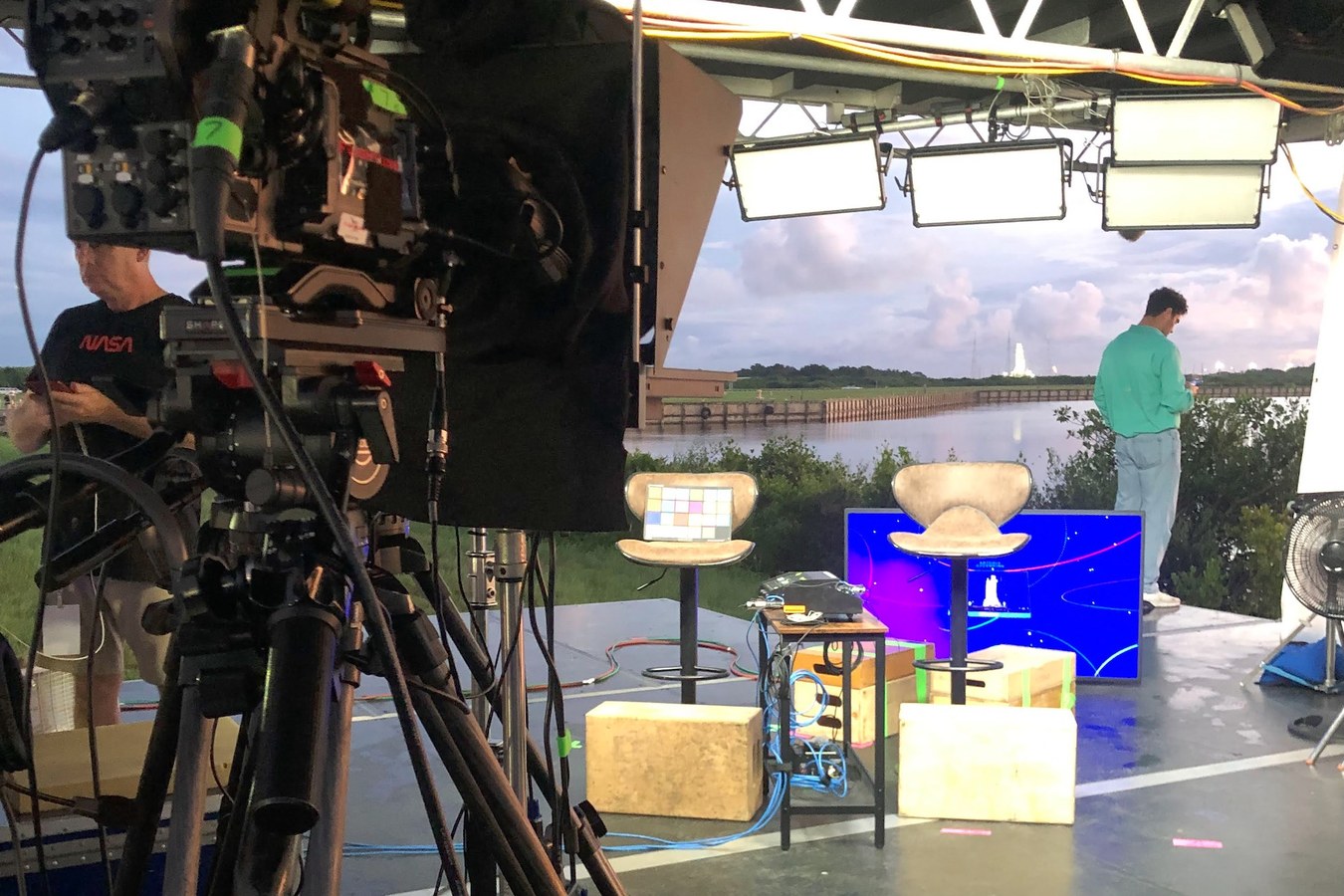 Broadcast camera and lit interview set beside the water at Kennedy Space Center, with the Artemis rocket on the pad in the distance